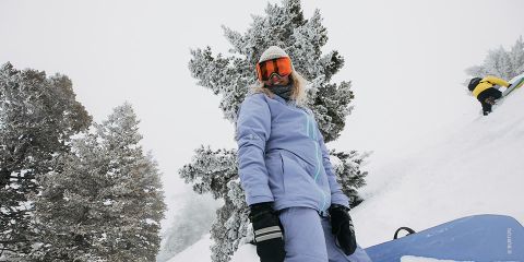 Ein Snowboarder in hellblauem Outfit und orangefarbener Schutzbrille steht auf einem schneebedeckten Hang und hält ein Snowboard. Im Hintergrund sind schneebedeckte Bäume zu sehen und eine weitere Person in einer gelben Jacke ist zu sehen, die den Hügel hinaufklettert.