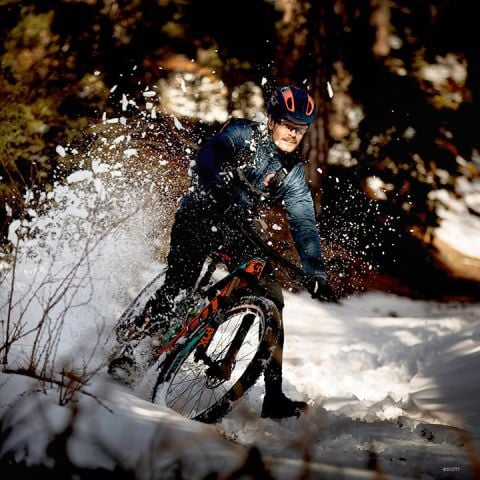 Eine Person auf einem Mountainbike rutscht durch verschneites Gelände und wirbelt dabei Schnee auf. Sie trägt einen Helm und Winterkleidung. Die Szene spielt in einem Waldgebiet mit verschwommenen Bäumen im Hintergrund.