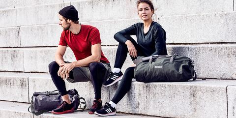 Ein Mann und eine Frau in Sportkleidung sitzen auf Betonstufen im Freien. Der Mann trägt ein rotes Hemd, Shorts und schwarze Leggings, während die Frau ein dunkles Oberteil und Leggings trägt. Beide haben Reisetaschen neben sich.