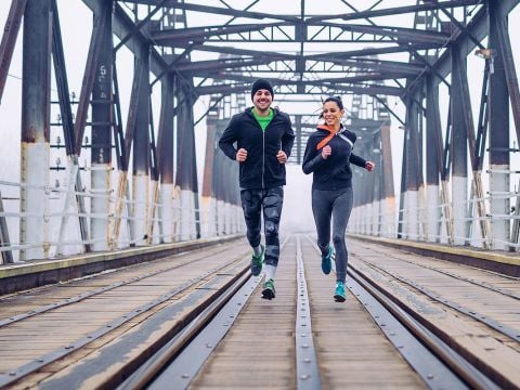 Zwei Menschen joggen nebeneinander über eine Holzbrücke mit Metallträgern. Sie tragen Sportkleidung und lächeln. Durch die Brückenkonstruktion ist ein nebliger Hintergrund zu sehen.