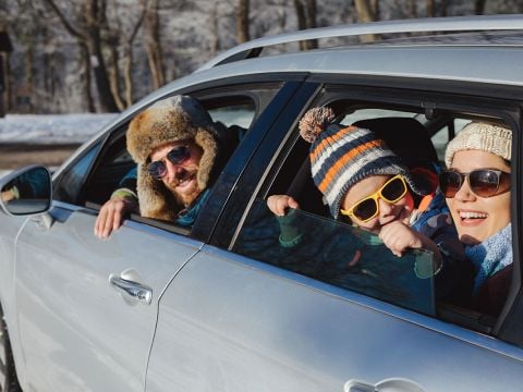 Eine Familie in Winterkleidung lehnt lächelnd aus einem silbernen Auto. Der Mann trägt eine Pelzmütze, das Kind eine Beanie und eine Sonnenbrille und die Frau eine Strickmütze und eine Sonnenbrille. Im Hintergrund sind schneebedeckte Bäume zu sehen.