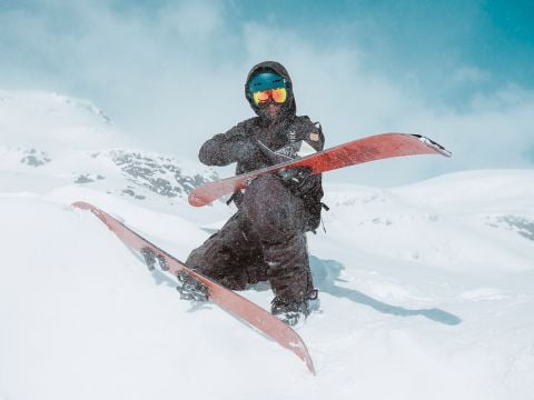 Eine Person in schwarzer Skiausrüstung hält gekreuzte Skier und kniet im Schnee. Die Skibrille spiegelt die verschneite Landschaft wider. Im Hintergrund sind schneebedeckte Berge und ein blauer Himmel zu sehen, die eine ruhige Alpenszenerie schaffen.