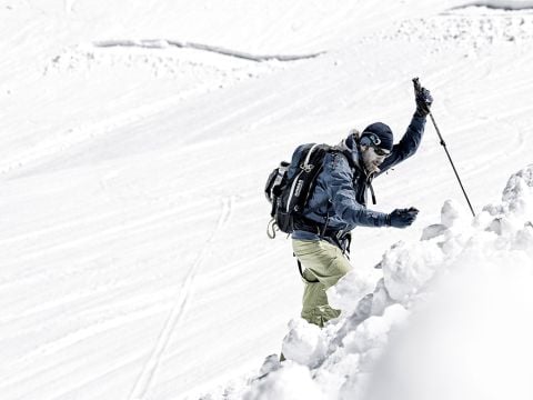 Eine Person in Winterkleidung erklimmt einen schneebedeckten Berghang und stützt sich dabei auf einen Stock. Im Hintergrund ist eine weite Schneefläche zu sehen, der Himmel ist nicht sichtbar. Die Szene vermittelt ein Gefühl von Abenteuer und Entschlossenheit.
