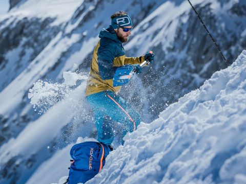 Eine Person in Winterkleidung und Sonnenbrille schaufelt Schnee auf einem schneebedeckten Berghang. Sie ist umgeben von sich bewegenden Schneeflocken und schroffen Berggipfeln im Hintergrund. Die Szene ist dynamisch und actionreich.