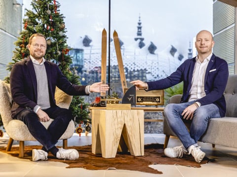 Zwei Männer sitzen in einem Innenraum mit einem Weihnachtsbaum auf der linken Seite. Sie lächeln und zeigen auf einen Tisch mit einer Holzauszeichnung und einem Retro-Radio. Große Fenster geben den Blick auf eine verschneite Stadtlandschaft im Hintergrund frei.