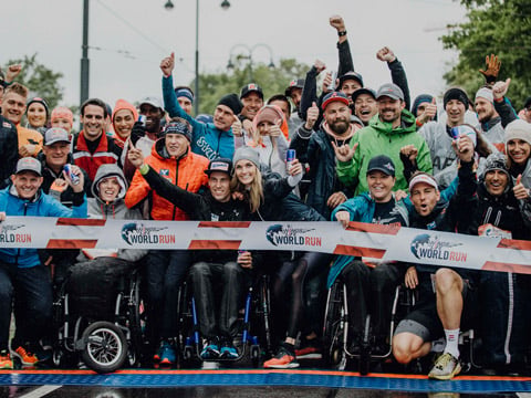 Eine bunt gemischte Gruppe von Menschen posiert aufgeregt an der Startlinie eines Laufevents. Einige sitzen im Rollstuhl. Sie tragen Sportkleidung und halten große Energydrink-Dosen in den Händen. Vor der Gruppe ist ein Banner mit der Aufschrift „World Run“ zu sehen.