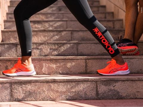 Eine Person trägt schwarze Leggings mit rotem „Tokio“-Aufdruck und leuchtend orangefarbene Laufschuhe und macht Dehnübungen auf Steinstufen. Eine andere Person in Shorts und passenden Schuhen steht in der Nähe und präsentiert Sportschuhe und Sportausrüstung.