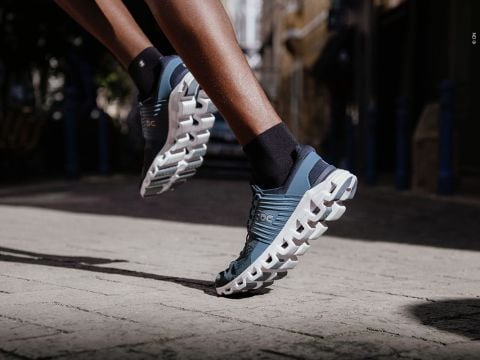 Nahaufnahme einer Person, die auf einer Straße läuft. Sie trägt blau-weiße Sportschuhe mit einzigartigen, wolkenartigen Sohlen. Der Fokus liegt auf dem Schuhwerk, das den Läufer mitten im Schritt einfängt und Bewegung und Beweglichkeit hervorhebt.