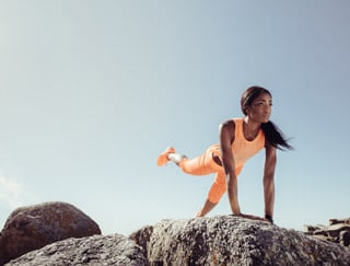 Eine Frau in leuchtend orangefarbener Sportkleidung balanciert auf Felsen und macht dabei eine Plankenübung, wobei sie ein Bein nach hinten streckt. Der Himmel ist klar und blau, was auf einen sonnigen Tag schließen lässt.