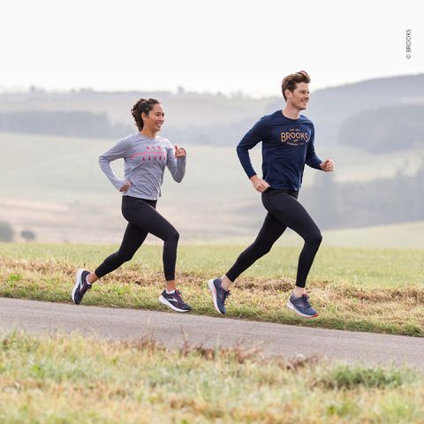 Zwei Personen joggen auf einem gepflasterten Weg in einer idyllischen Landschaft. Die Frau trägt ein graues Langarmshirt und schwarze Leggings, während der Mann ein dunkelblaues Langarmshirt und schwarze Leggings trägt. Beide tragen Laufschuhe. Im Hintergrund sind Felder und Bäume zu sehen.