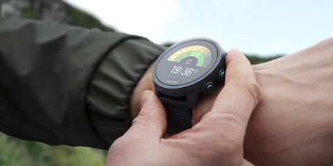 Nahaufnahme einer Person, die im Freien eine schwarze Smartwatch an ihrem Handgelenk einstellt. Das Zifferblatt zeigt die digitale Zeit 19:36:21 an und verfügt über ein Kompassdesign mit einem gelb-grünen Farbverlauf. Die Person trägt eine grüne Jacke.