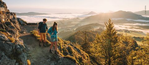 Zwei Menschen wandern bei Sonnenaufgang mit Rucksäcken und Wanderausrüstung einen felsigen Bergpfad entlang. Unter ihnen erstreckt sich ein nebelverhangenes Tal und in der Ferne Hügel unter einem goldenen, sonnendurchfluteten Himmel. Üppiges Grün umgibt die Gegend.