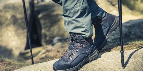 Eine Person in dunklen Wanderschuhen und grünen Hosen steht mit Trekkingstöcken auf einem Felsen. Im Hintergrund sind verschwommene Felsen und Blätter zu sehen. Das Bild fängt einen Moment während einer Wanderung in einer natürlichen Umgebung ein.