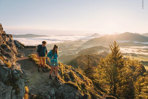 Zwei Wanderer, ein Mann und eine Frau, gehen einen felsigen Pfad entlang, von dem aus man ein Tal überblicken kann. Die Sonne geht hinter den Bergen in der Ferne unter und wirft einen warmen Schein auf eine Landschaft mit sanften Hügeln und vereinzelten Wolken. Unten sind Bäume und grüne Felder zu sehen.