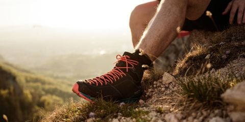 Eine Person in Wanderschuhen mit leuchtend orangefarbenen Schnürsenkeln sitzt auf einem felsigen Untergrund. Ihr Bein ist ausgestreckt, im Hintergrund bietet sich ein malerischer Blick auf ein sonnenbeschienenes Tal und entfernte Berge. Sie ist von Gras und kleinen Steinen umgeben.