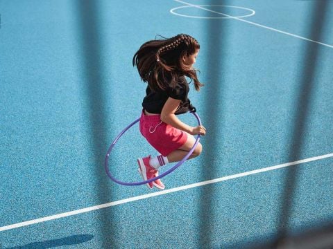 Ein junges Mädchen mit geflochtenen Haaren springt auf einem blauen Sportplatz durch einen lila Hula-Hoop-Reifen. Sie trägt ein schwarzes Shirt, rosa Shorts und rote Turnschuhe mit weißen Socken. Das Bild ist teilweise von unscharfen vertikalen Linien eingerahmt.