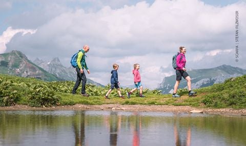 Eine vierköpfige Familie wandert auf einem malerischen, grasbewachsenen Pfad mit einem ruhigen Teich im Vordergrund. Im Hintergrund sind Berge unter einem teilweise bewölkten Himmel zu sehen. Jedes Mitglied trägt farbenfrohe Wanderkleidung und einen Rucksack.