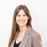 Lächelnde Frau mit langen braunen Haaren, trägt einen karierten Blazer und ein schwarzes Oberteil und steht vor einem schlichten weißen Hintergrund.