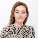 Eine Frau mit braunen Haaren lächelt in die Kamera. Sie trägt einen beigen Pullover mit schwarzem Zebrastreifenmuster. Der Hintergrund ist schlicht weiß.