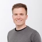 Lächelnde Person mit kurzen Haaren und grauem Pullover, die vor einem schlichten weißen Hintergrund steht.