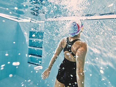 Ein Schwimmer in einem schwarzen Badeanzug mit weißer Badekappe liegt unter Wasser in einem Pool. Er ist von Blasen umgeben und blickt nach oben, während Sonnenlicht durch das Wasser fällt. Im Hintergrund ist eine geflieste Wand zu sehen.