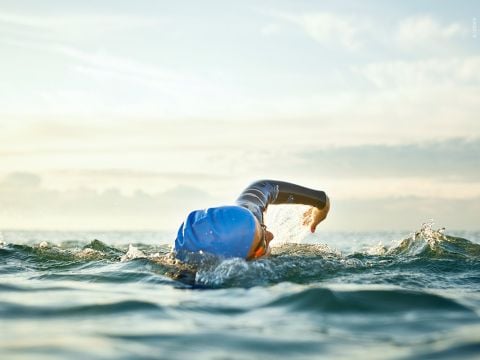 Ein Schwimmer mit blauer Badekappe und Schwimmbrille schwimmt im offenen Wasser Kraulstil. Die sonnenbeschienenen Wellen plätschern um sie herum und der Horizont ist unter einem sanften, bewölkten Himmel sichtbar.