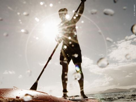Eine Person fährt Stand-Up-Paddleboard auf dem Meer. Die Silhouette ist vor einer hellen Sonne zu sehen, im Vordergrund sind Wassertropfen zu sehen. Der Himmel ist teilweise bewölkt und am Horizont sind in der Ferne Berge zu sehen.