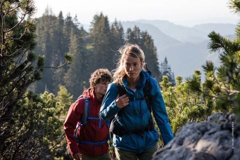 Zwei Personen wandern einen felsigen Pfad hinauf, der von üppigen grünen Bäumen umgeben ist. Die Person vorn trägt eine blaue Jacke, während die Person dahinter eine rote Jacke trägt. Im Hintergrund sind Bergkämme unter einem klaren Himmel zu sehen.
