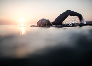 Ein Schwimmer mit Badekappe schwimmt bei Sonnenuntergang im offenen Wasser Kraulschwimmen. Die Sonne wirft einen warmen Schein auf den Horizont und durch den Schwimmzug des Schwimmers spritzen Wassertropfen in die Luft. Die Szene ist ruhig und heiter.