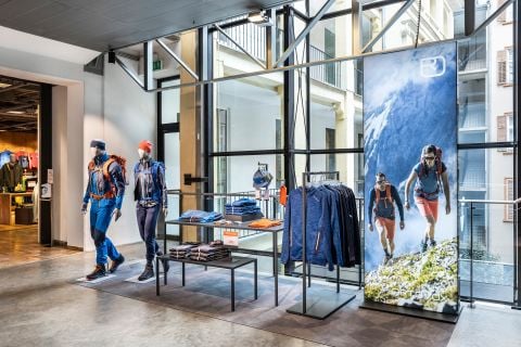Schaufenster mit Outdoor-Wanderpuppen in blauer und orangefarbener Kleidung. Die Kleidung ist ordentlich auf Ständern und Tischen angeordnet. Großes Fenster im Hintergrund. Auffälliges Poster mit Wanderern in einer Berglandschaft auf der rechten Seite.