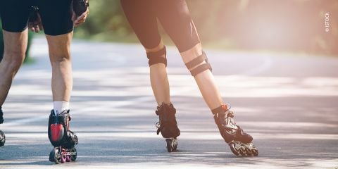 Zwei Personen fahren auf Rollschuhen auf einem glatten Weg, umgeben von Grün. Der Fokus liegt auf ihren Unterkörpern, die Sportbekleidung und Rollschuhe zeigen. Sonnenlicht wirft weiche Schatten auf den Bürgersteig.