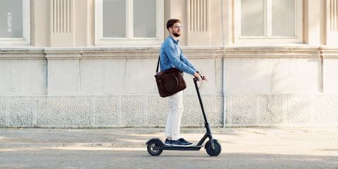 Eine Person mit Bart und Brille fährt mit einem Elektroroller eine Straße entlang. Sie trägt ein blaues Hemd, weiße Hosen und eine braune Tasche. Sie kommt an einem Gebäude mit großen Fenstern und einer beigen Fassade vorbei.
