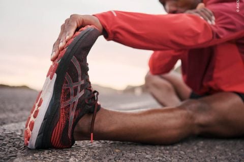 Eine Person in einer roten Jacke streckt ihre Beine auf dem Bürgersteig aus. Sie trägt schwarze und rote Sportschuhe. Der Hintergrund ist unscharf, sodass die Dehnungsbewegung und das Schuhwerk im Vordergrund stehen.