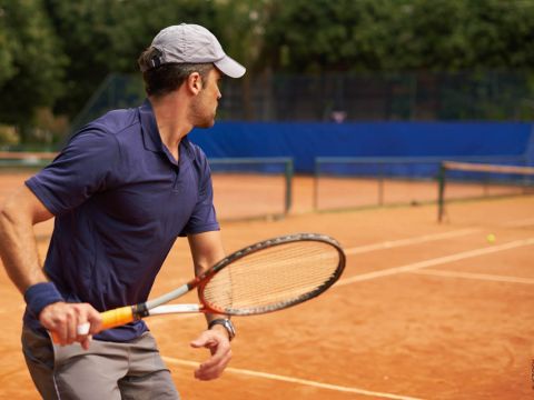 Eine Person in blauem Hemd und grauer Mütze spielt Tennis auf einem Sandplatz. Sie hält einen Tennisschläger in der Hand und scheint sich auf das Spiel zu konzentrieren. Im Hintergrund sind ein Netz und grüne Bäume zu sehen.