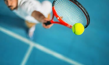 Eine Person wird dabei gefilmt, wie sie einen Tennisball auf einem blauen Platz aufschlägt. Sie hält einen rot-schwarzen Tennisschläger. Der Fokus liegt auf Ball und Schläger, die leicht verschwommene Bewegung lässt auf einen schnellen Aufschlag schließen.