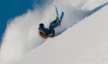 Ein Skifahrer mit blauer Jacke und schwarzem Helm stürzt auf einem schneebedeckten Hang, die Skier sind in der Luft und werden von einer dicken Schneewolke umhüllt. Im Hintergrund ist ein klarer blauer Himmel zu sehen.