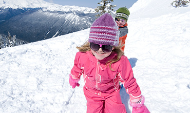 Zwei Kinder spielen an einem sonnigen Tag im Schnee und tragen bunte Winterkleidung. Das Mädchen vorne trägt einen rosa Schneeanzug mit einer lila Mütze und der Junge dahinter ein orangefarbenes Outfit mit einer grünen Mütze. Im Hintergrund sind schneebedeckte Berge zu sehen.