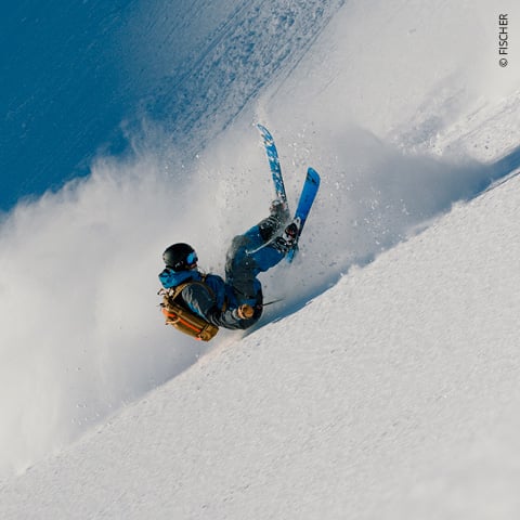 Ein Skifahrer in blauer Jacke und Helm fährt einen verschneiten Hang hinunter und wirbelt dabei eine Schneewolke auf. Der Skifahrer neigt sich zur Seite, im Hintergrund ist der blaue Himmel zu sehen.