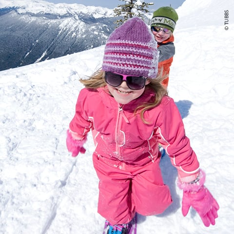 Zwei Kinder in Winterkleidung spielen an einem sonnigen Tag im Schnee. Das vordere Kind trägt einen rosa Schneeanzug, eine gestreifte Mütze und eine Sonnenbrille. Das Kind dahinter ist orange und grün gekleidet. Im Hintergrund sind schneebedeckte Berge zu sehen.