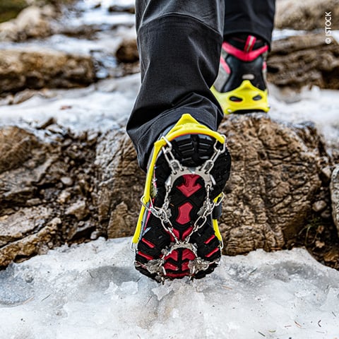 Nahaufnahme einer Person mit Eiskrallen an bunten Wanderschuhen, die über felsiges, eisiges Gelände läuft. Der Fokus liegt auf den Krallen, die auf der rutschigen Oberfläche Halt finden und zeigt die Ausrüstung für Outdoor-Winteraktivitäten.