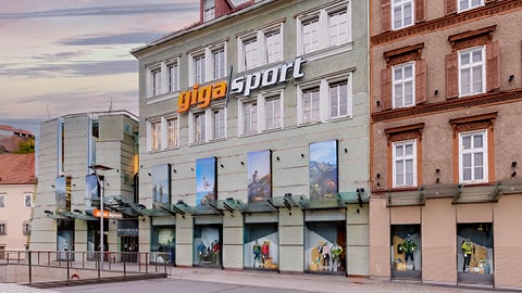 Straßenansicht eines Gigasport-Geschäfts in einem alten Gebäude im europäischen Stil. Die Fassade ist hellgrün und braun und hat mehrere Fenster. Große Ladenschilder sind sichtbar und in den Fenstern werden verschiedene Außenbilder angezeigt.