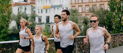 Eine Gruppe von fünf Personen joggt über eine mit Liebesschlössern geschmückte Brücke. Sie tragen passende weiße Sportoberteile und wirken konzentriert. Im Hintergrund sind Bäume und Gebäude zu sehen, darunter eines mit einem „Giga Sport“-Schild.