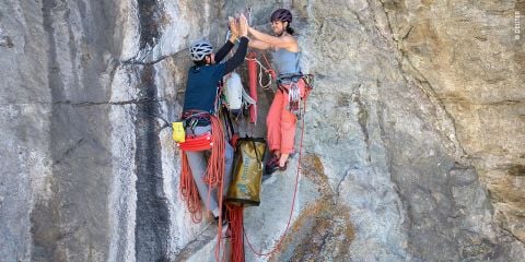 Zwei Kletterer mit Helmen geben sich an einer Felswand ein High-Five. Sie sind mit Seilen gesichert und ihre Kletterausrüstung ist an Gurten befestigt. Im Hintergrund sind schroffe, strukturierte Felsoberflächen zu sehen.