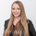 Frau mit langen blonden Haaren, trägt eine schwarze Lederjacke und ein schwarzes Oberteil und lächelt vor einem schlichten weißen Hintergrund.