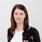 Eine Frau mit braunem Haar trägt eine schwarze Jacke mit Blumenmuster über einem weißen Oberteil. Sie blickt mit einem leichten Lächeln in die Kamera. Der Hintergrund ist schlicht und hell.