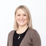 Eine Frau mit schulterlangem blondem Haar und einem Lächeln trägt ein schwarzes Oberteil und eine braune Strickjacke. Sie trägt eine Perlenkette und steht vor einem schlichten weißen Hintergrund.