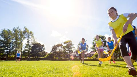 Eine Gruppe Kinder spielt Feldhockey auf einem Rasenplatz. Sie tragen Sportkleidung und halten gelbe Hockeyschläger in der Hand. Die Sonne scheint hell, im Hintergrund sind Bäume und Häuser unter einem klaren Himmel zu sehen.