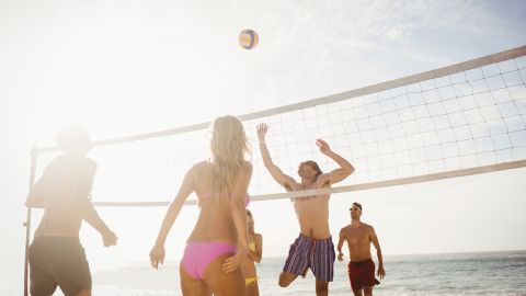 Menschen spielen Beachvolleyball unter einem sonnigen Himmel. Eine Frau in rosa Badebekleidung und ein Mann in gestreiften Shorts springen in der Nähe des Netzes, um den Ball zu schlagen, während andere zuschauen und sich auf das Spiel vorbereiten. Im Hintergrund ist das Meer zu sehen.