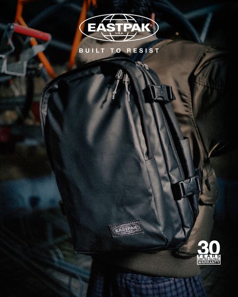 Eine Person in einer braunen Jacke trägt einen schwarzen Eastpak-Rucksack mit dem Slogan „Built to Resist“. Das Bild zeigt ein Logo und die Erwähnung einer „30-jährigen Garantie“. Der Hintergrund ist unscharf.