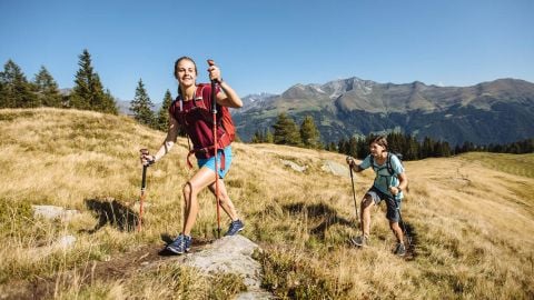 Zwei Personen wandern unter einem klaren blauen Himmel auf einem Graspfad in den Bergen. Die Frau vorn lächelt, trägt einen Rucksack und benutzt Wanderstöcke. Ein Mann folgt ihnen, ebenfalls mit Rucksack und Stöcken. In der Ferne sind Berggipfel zu sehen.